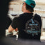 A man wearing a turquoise cap and a black cotton t-shirt with a graphic wizard hat, wolf, and the text “Howler Brothers” sits outdoors at a picnic table, facing away from the camera, capturing a subtle vintage look.