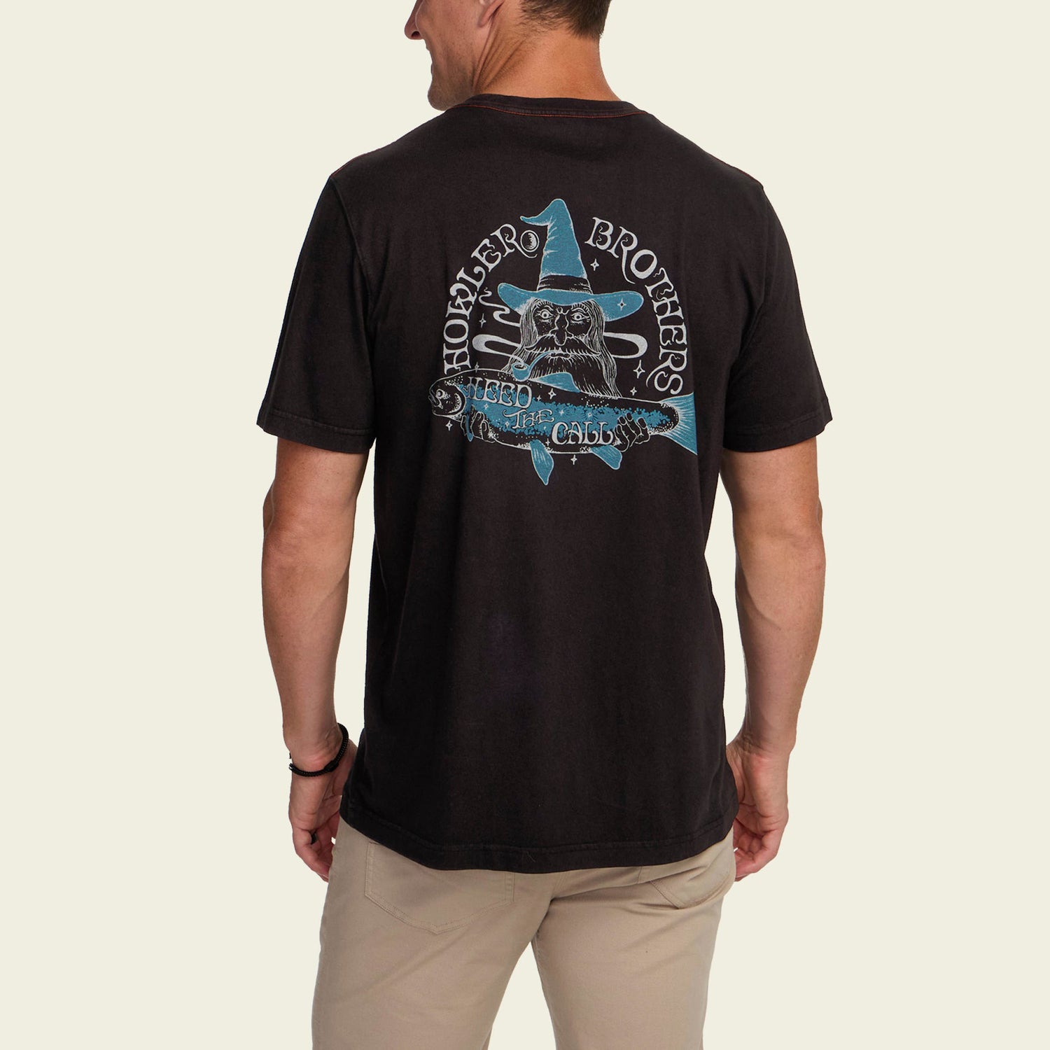 A man wearing a black cotton T-shirt with a vintage look, featuring experimental artwork of a wizard, blue star, and the text Hopper Bros and Need Chaos on the back. He stands facing away in beige pants.