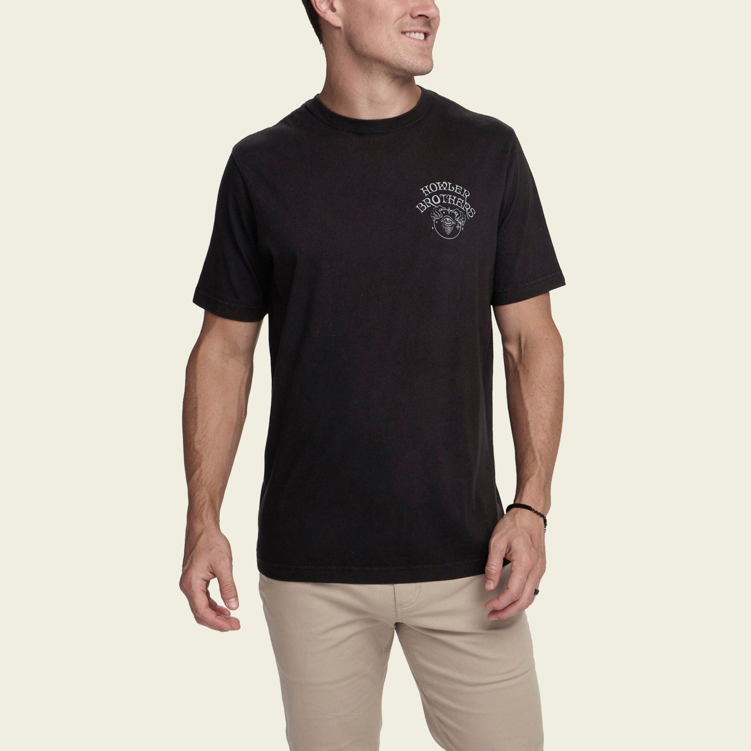 A man wearing a black cotton T-shirt with a small white Howler Brothers logo on the left chest and light-colored pants stands against a plain light background, showcasing a subtle vintage look.