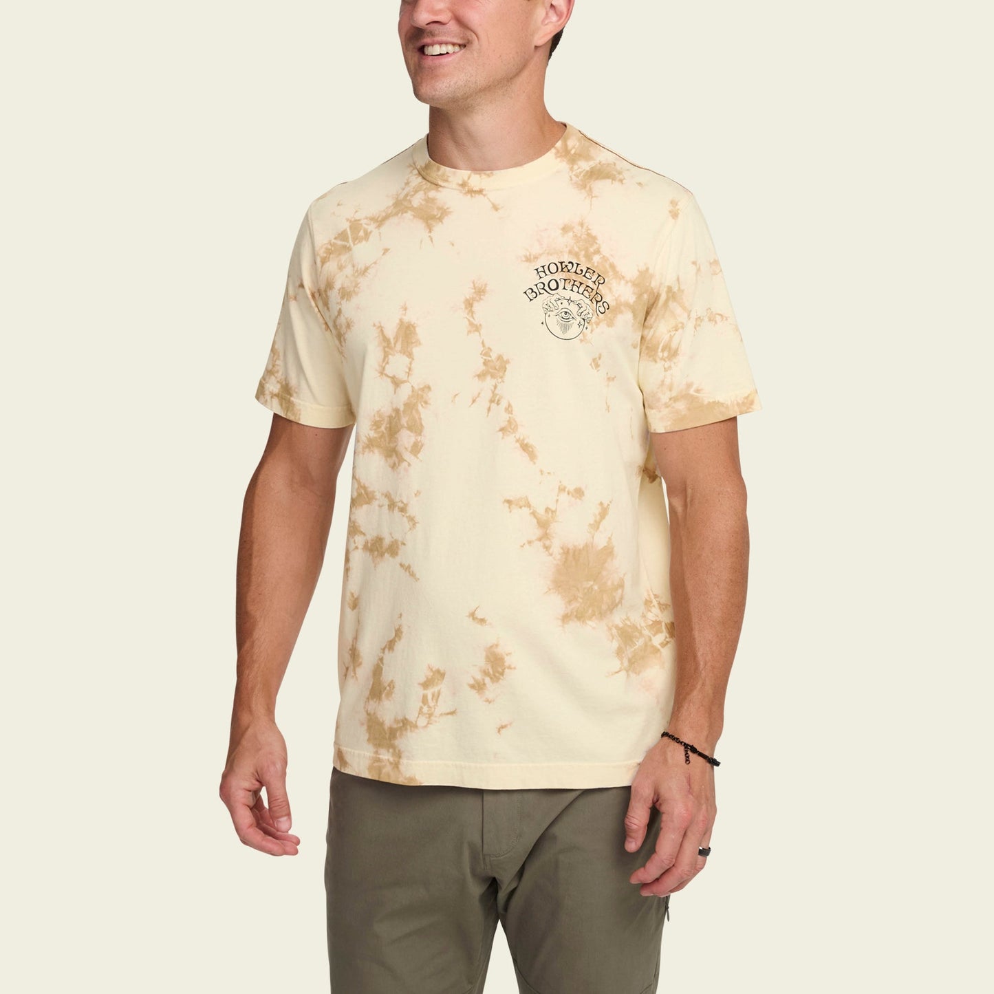 A person wearing a light beige and brown tie-dye cotton T-shirt with a small logo and text on the left chest, paired with olive green pants, stands against a plain background, smiling with arms relaxed for a subtle vintage look.