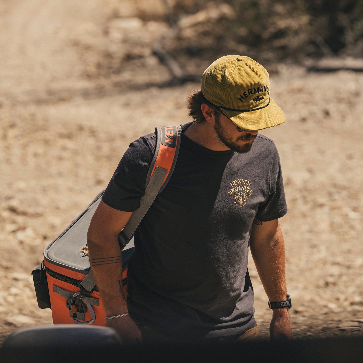 A man in a yellow cap, sunglasses, and a black cotton-poly blend t-shirt carries an orange cooler bag over his shoulder while walking outdoors on a dirt path in sunny, dry conditions.