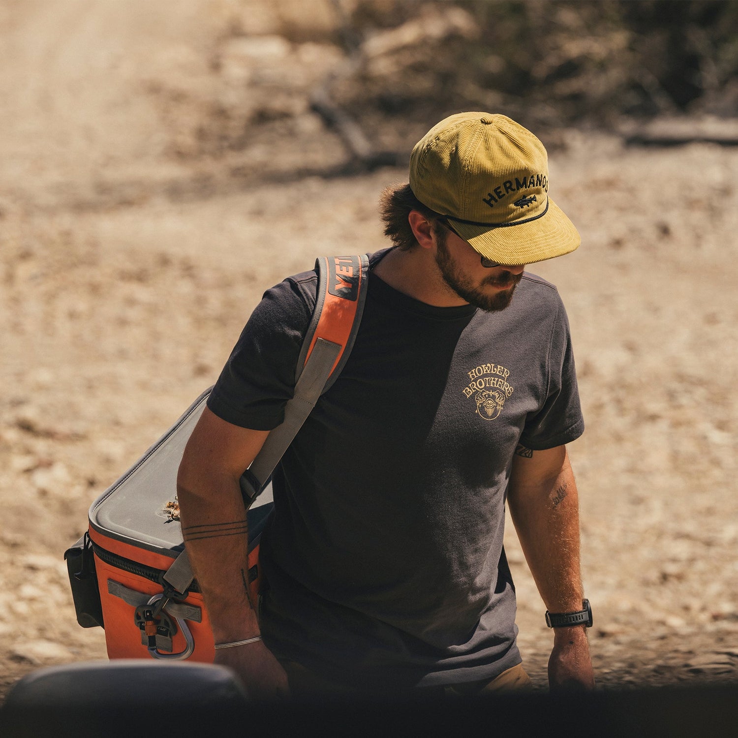 A man in a yellow cap, sunglasses, and a black cotton-poly blend t-shirt carries an orange cooler bag over his shoulder while walking outdoors on a dirt path in sunny, dry conditions.