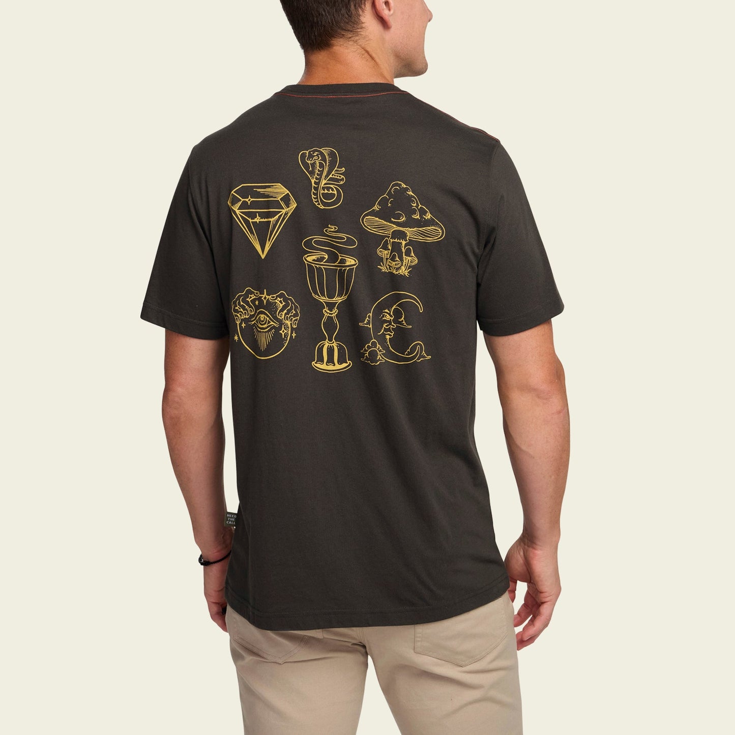 A man wears a black Howler t-shirt with gold line art—diamond, serpent, mountain, heart, chalice, crescent moon, and a hand with an eye. The cotton-poly blend ensures comfortable t-shirts while showcasing bold back artwork.