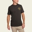 A man wearing a black Howler t-shirt with a small yellow logo on the left chest stands smiling in light khaki pants against an off-white background, showing off the comfort of these cotton-poly blend comfortable t-shirts.