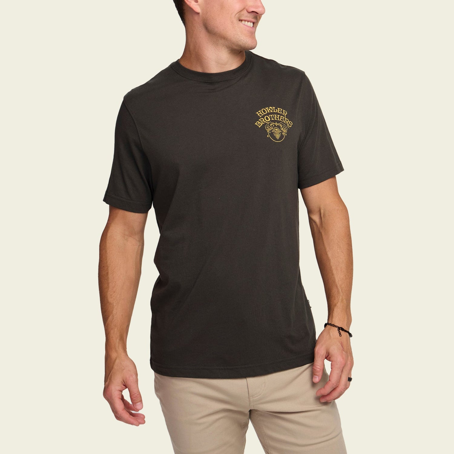 A man wearing a black Howler t-shirt with a small yellow logo on the left chest stands smiling in light khaki pants against an off-white background, showing off the comfort of these cotton-poly blend comfortable t-shirts.