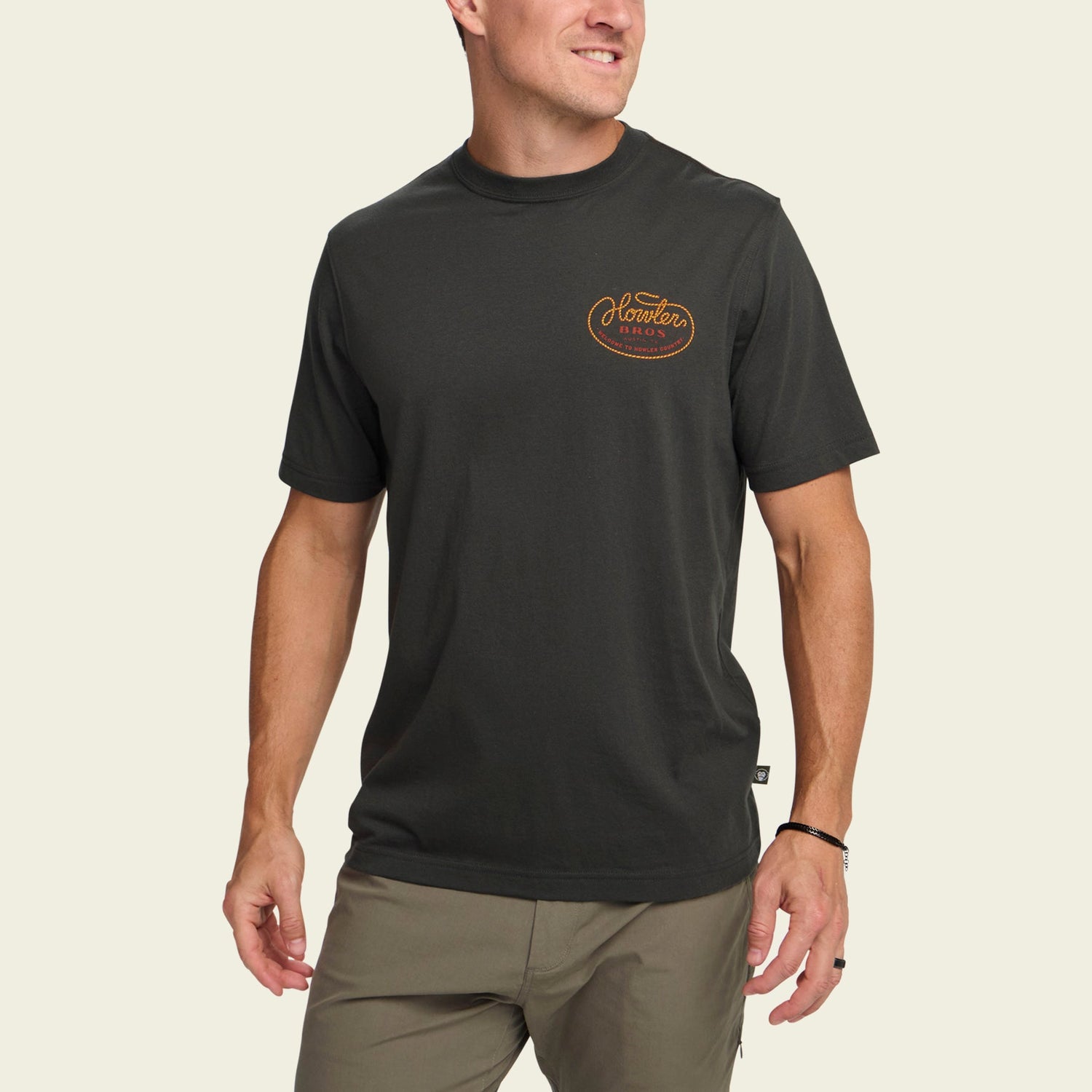 A man wearing a black short-sleeve t-shirt made from a soft cotton-poly blend with an orange Outdoor Research logo on the left chest, paired with olive green pants, stands against a plain light background and smiles while looking to the side.