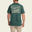 A man wearing a green Howler blended t-shirt with a large cowboy graphic on the back, made from a cotton-poly blend for maximum comfort. He pairs it with light-colored pants and faces away from the camera.