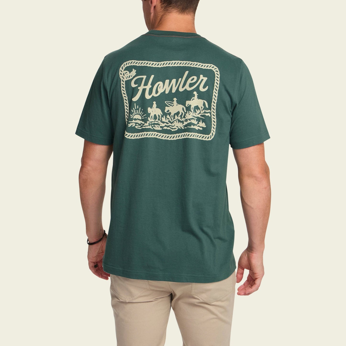 A man wearing a green Howler blended t-shirt with a large cowboy graphic on the back, made from a cotton-poly blend for maximum comfort. He pairs it with light-colored pants and faces away from the camera.