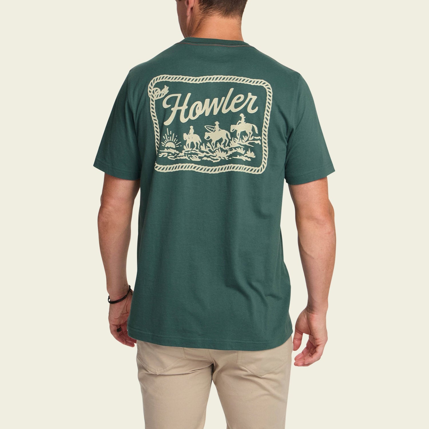 A man wearing a green Howler blended t-shirt with a large cowboy graphic on the back, made from a cotton-poly blend for maximum comfort. He pairs it with light-colored pants and faces away from the camera.