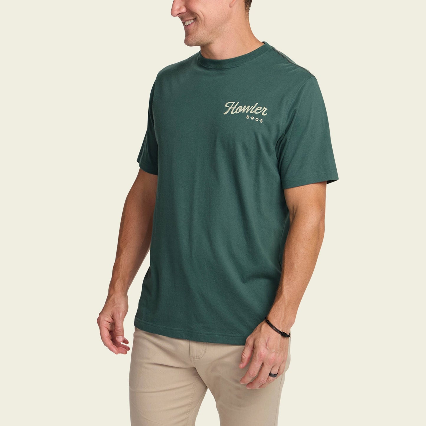 A man wearing a dark green Howler blended t-shirt with Howler Bros on the chest and beige pants stands smiling against an off-white background, showcasing maximum comfort and style.