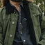Close-up of a person wearing an olive green corduroy jacket with a black sherpa collar over a dark charcoal button-up shirt. A small chest patch on the shirt reads 'SHEER'.