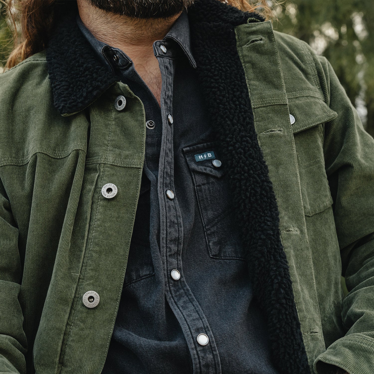 Close-up of a person wearing an olive green corduroy jacket with a black sherpa collar over a dark charcoal button-up shirt. A small chest patch on the shirt reads 'SHEER'. - View 6