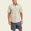 A man wearing a lightweight cotton, patterned single pocket shirt with blue shorts stands and smiles against a plain light background.