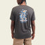 A person wearing a dark gray Howler blended T-shirt with a colorful graphic and the words HOWLER HEED THE CALL on the back, standing with their back to the camera and sporting light beige shorts for maximum comfort.