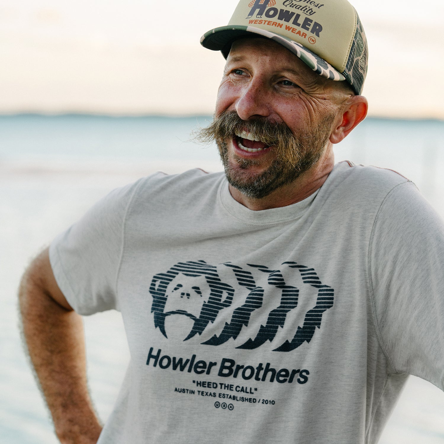 Man wearing a gray 'Howler Brothers' t-shirt and cap by the water