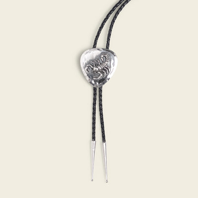 The El Alacrán Bolo Tie by Cityboy Forge features a heart-shaped silver slide with a raised design and two black braided cords ending in silver tips, beautifully showcasing this handmade bolo tie on a plain light background.